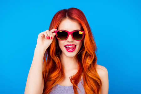 Funny carefree girl with ginger hair holding sunglasses and licking pink lips while standing on blue backgroundの写真素材