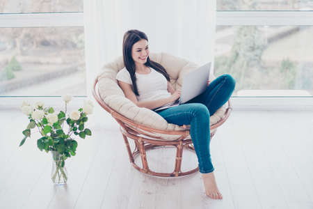 Cheerful young cute pretty woman enjoying  weekend while sitting on comfort soft modern armchair with white rose at homeの写真素材