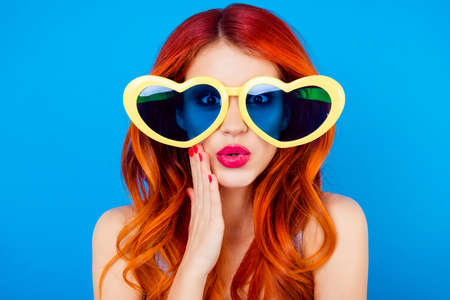 Portrait of carefree cheerful pretty trendy charming giri with curly hair in shock, she is fall in love while touch face and look through glasses like heart form at camera  on blue backgroundの写真素材