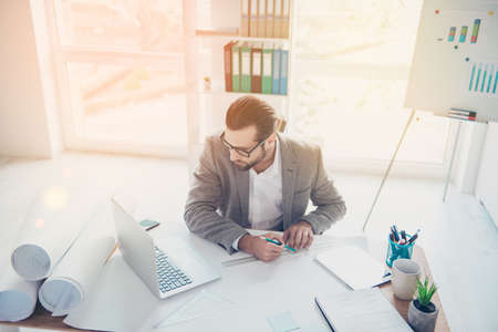 Top-view of handsome concentrated man in formal wear working with blueprint papers and drawings in modern lightful officeの写真素材