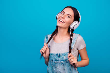 Portrait of cute young girl with funny pigtails in jeans overalls listening to music against blue backgroundの写真素材