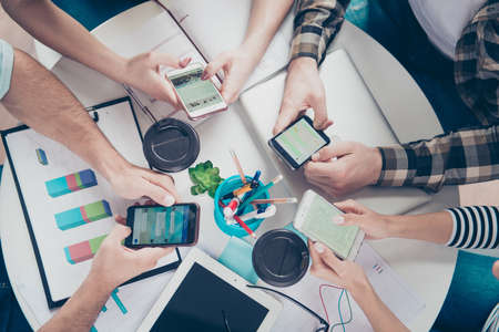 Top up of four friends using their smartphones for searching ideas for new successful business in work environment sitting by the tableの写真素材