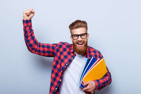 Young cheerful student in glasses and in checkered shirt with books in hands celebrating ending of exams