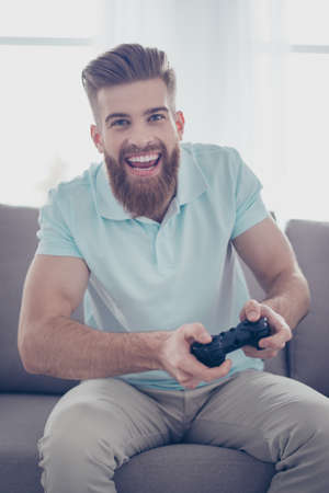 A portrait of young handsome bearded man holding joystick and playing videogames.He sits at home on...