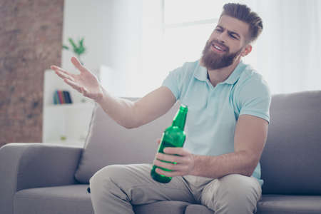 Upset young man is frustrated about the loss of the team in game he is watching. Guy is having beer and sitting on the sofa at livingroomの写真素材