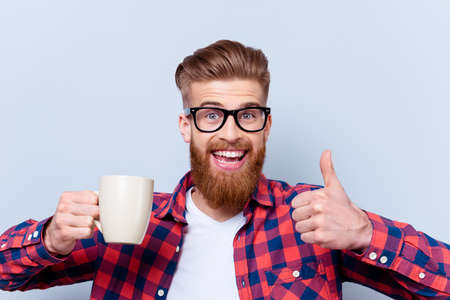 Close up photo of crazy happy smiling man in spectacles holding cup of tasty fresh coffee and showing thumb upの写真素材