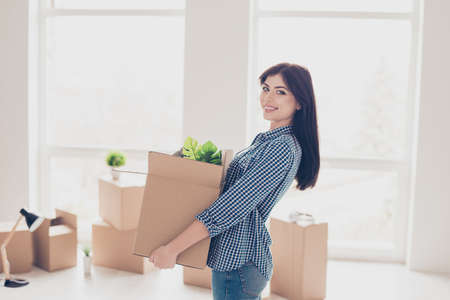 Young cheerful successful woman moving to new nice place and holding box with her belongings. The room is very light and bright, a lot of boxes with stuff around herの写真素材