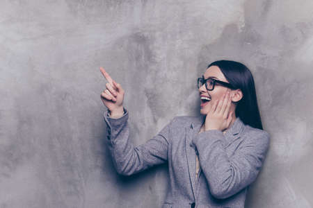 Portrait of excited cheerful young successful businesswoman in glasses and suit pointing to the side  while standing near gray wallの写真素材