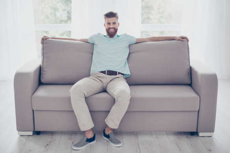 Portrait of relaxed smiling handsome bearded man resting on cozy sofa at living room at homeの写真素材