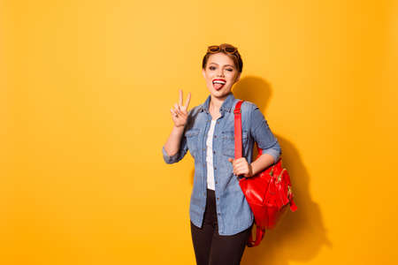 Playful young girl with red lipstick and tongue out in stylish sunglasses on the yellow background. She is excited, holding her bright red backpackの写真素材