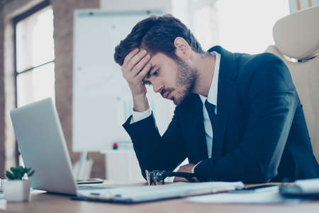 Young tired, ill, overworked  man in formal wear sitting in front of computer and touching his foreheadの写真素材