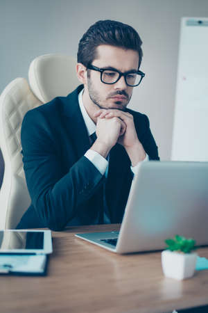 Vertical portrait of minded entrepreneur  in spectacles holding his hands under chin and reading something from computerの写真素材