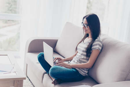 Happy smiling lady in spectacles sitting on sofa and using computer for her workの写真素材
