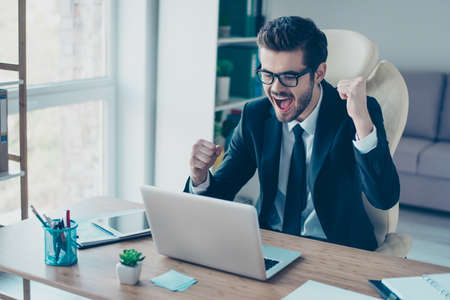 Portrait of happy screaming young businessman in formal-wear with tie  sitting at the computer and triumphing with raised fists because of successful resultsの写真素材