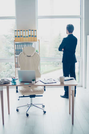Vertical photo of minded man in suit standing with crossed arms and looking through the window of his modern officeの写真素材