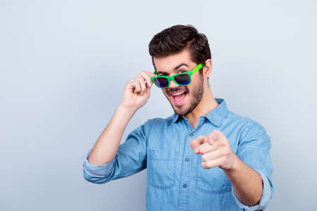cool playful handsome young man in bright green glasses is pointing on camera with open mouthの写真素材