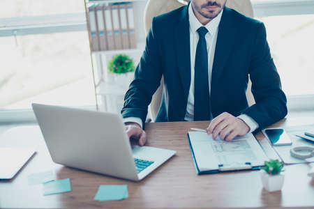 Close up cropped photo of concentrated minded businessman in black and white formal wear with tie sitting at the table with laptop and studing the report in detailの写真素材