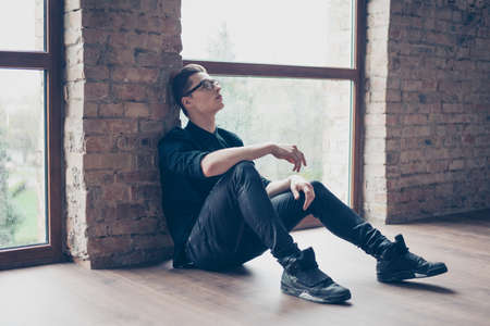 Young cute pensive boy is looking up and sitting on the floor near the window. He is in black...