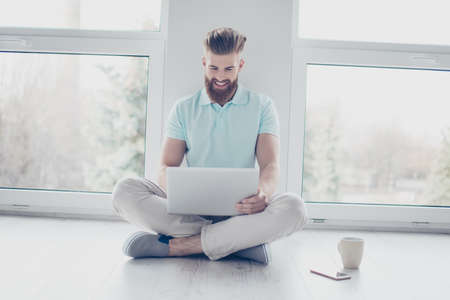 Cheerful student is studying with his laptop sitting on the floor at home. He feels relaxed and...