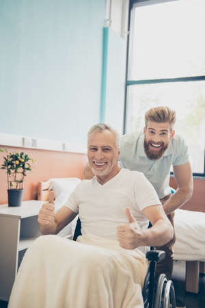 Yes! Grandpa got cured! Excited young bearded grandson is taking his granpa home in a wheel chair after recovery from the hospitalの写真素材