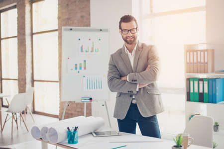 Confident smiling architect in formal wear and glasses standing with crossed hands at the table with papers and documents in modern officeの写真素材