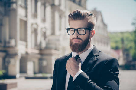 Close up portrait of a successful young red bearded guy in suit and glasses, fixing his tie. So stylish and nerdy. Outdoors on a sunny streetの写真素材