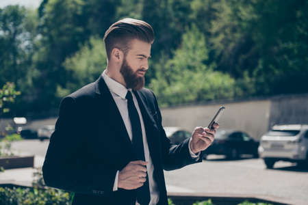 Young stylish bearded business man in a classy suit is browsing his pda on the street at the sunny day. He is ordering a cab by pdaの写真素材