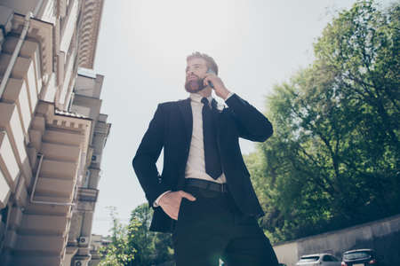 Low angle view of a young handsome bearded business man in a classy suit talking on his mobile outdoors at the sunny spring day. He looks stunningの写真素材