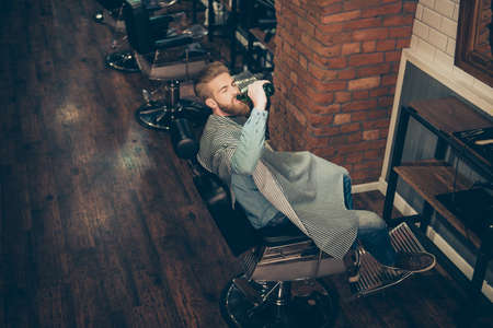 Chill out at the barber shop. Side view of handsome stylish young red bearded man drinking beer, while waiting to get a haircutの写真素材