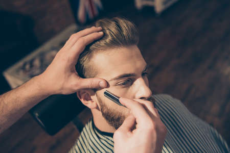 Close up of a hairdresser`s work for an attractive young blond man at the barber shop. He is doing styling of his beard, shaving it with straight razorの写真素材
