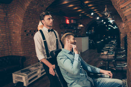 Young blond stylish guy in a casual jeans outfit is showing to barber shop worker how he wants his beard to be done. Classy dressed hairdresser is looking focusedの写真素材