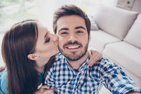 Attractive young couple is taking a selfie on camera, girl kisses her boyfriend, they are indoors at homeの写真素材