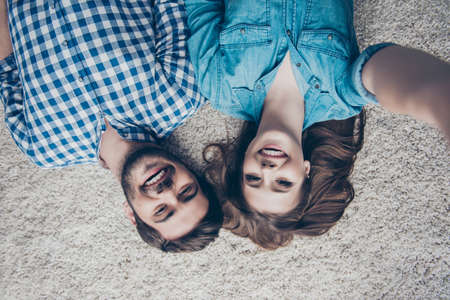 Upside down top view photo of two young friends, who are taking selfie and smiling. They lie on the floor on beige carpet in casual outfits indoors at homeの写真素材