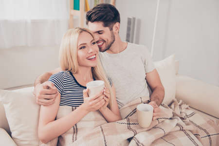 Cheerful romantic couple is sitting on sofa under cozy plaid and smiling. They are drinking tea, he is looking at her with tendernessの写真素材