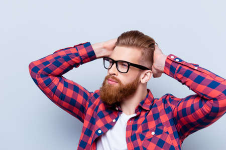 Stylish confident young bearded man in checkered shirt and glasses touching his haircut against gray...