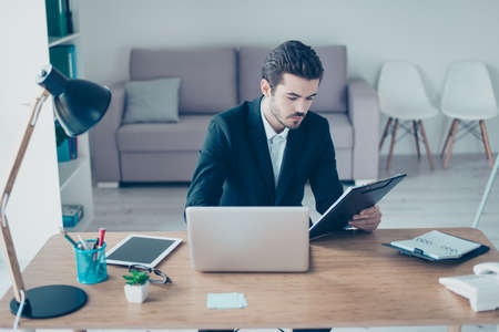 Portrait of successful young manager in formalwear using computer and clipboard for checking out the...