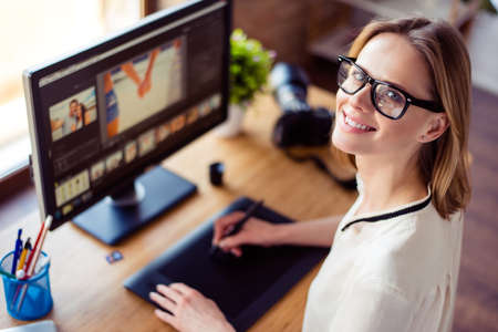 Top view of intelligent blond young woman working with computer and graphic tablet, stylus, smiling. She is a successful self employed retoucher and photographの写真素材