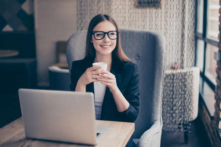 Time to make pause. Lunch time.  Dreamy inspired girl is sitting in the cafe and drinking tea, looking in the window, dressed in formal wear, glassesの写真素材