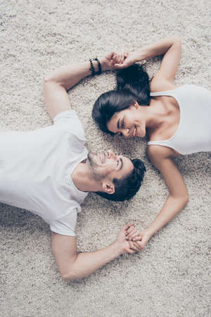 Look in my eyes forever. Dreamy couple of young beautiful lovers are lying on the beige cozy carpet indoors at home, holding each others hands. So sensualの写真素材