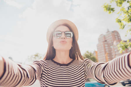 Selfie and an air kiss! Young cute lady is photographing outdoors in the summer city, wearing casual spring outfit, trendy hat and fashionable sunglassesの写真素材
