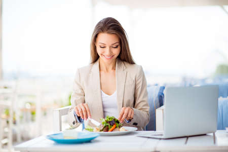 Break for energy, vitamins and refreshness. Young cheerful lady is having a business lunch on an open air light terrace outdoors. She is in a formal wear, having salad in front of laptopの写真素材