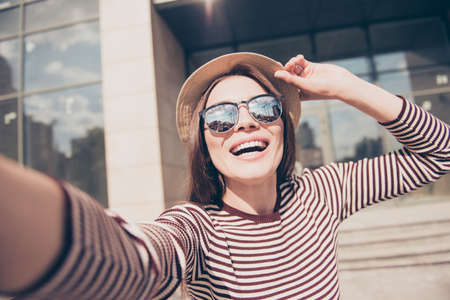 selfie mania excited young girl is making selfie on a camera. she is wearing casual striped spring outfit, sunglasses, fixing her cap, on a walk in town outdoorsの写真素材