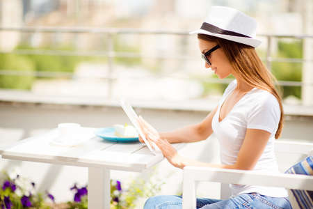 Profile portrait of young charming lady in casual outfit and hat, who is browsing news at her tablet with dessert and coffee in the light designed open air light terrace of summer cafeの写真素材