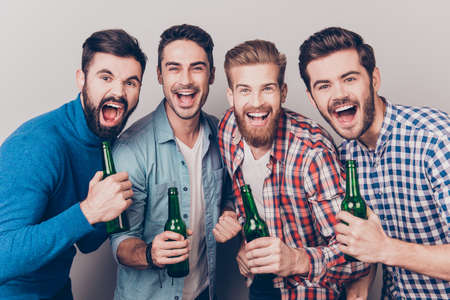 Men`s club. Four crazy friends guys are screaming with bottles in hands, all in casual shirts, isolated on grey background. They are fans of sports games as football, basketball, hockey, baseballの写真素材