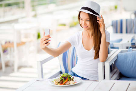 Young cheerful lady is having lunch on a summer open air light terrace outdoors. She is in a casual wear, having salad and taking selfie with her pda, fixing hatの写真素材