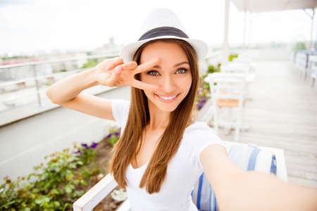 Cute young girl with toothy beaming smile is making selfie on a camera of her pda. She is wearing...