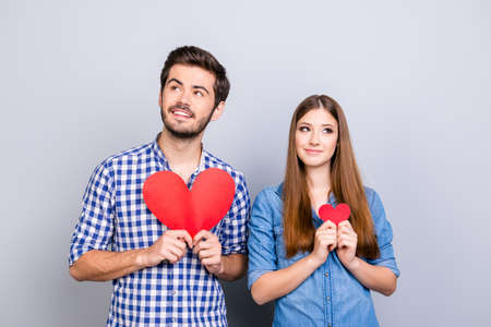 Two young cute lovers are looking away and smiling, holding big and small red paper hearts, wearing casual shirts, standing on the pure background near each otherの写真素材