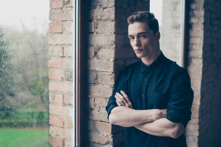 Young handsome man in black outfit is standing near the window with crossed arms and looking seriously in the cameraの写真素材