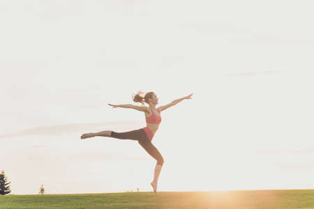Young gymnast in fashion sportswear is jumping in graceful pose on the sunset background, in a spring park outdoorsの写真素材