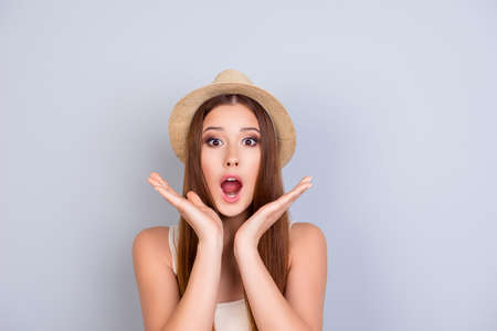 Shocked young attractive girl is holding her palms near her face. She is excited and with open mouth on pure light grey background, she is wearing summer beige hatの写真素材